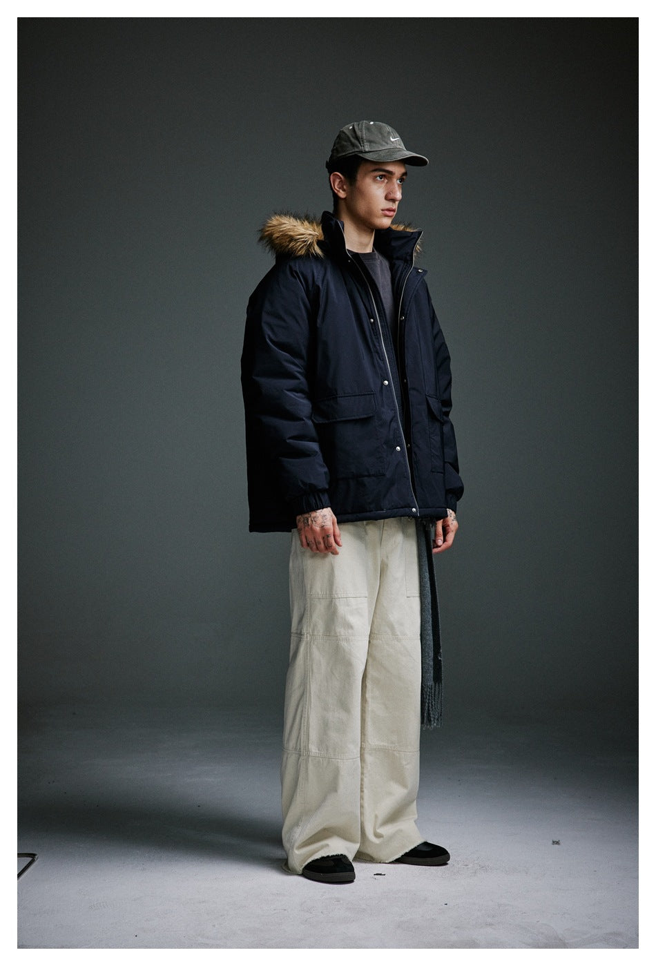 Model wearing a dark navy hooded jacket with faux fur-style trim over a dark tee, beige wide pants and a cap, front view