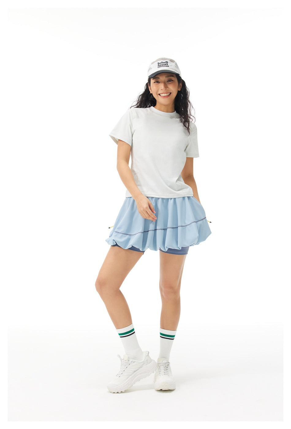 Model wearing a white short-sleeve top, pale blue flared tennis skirt, and white cap.