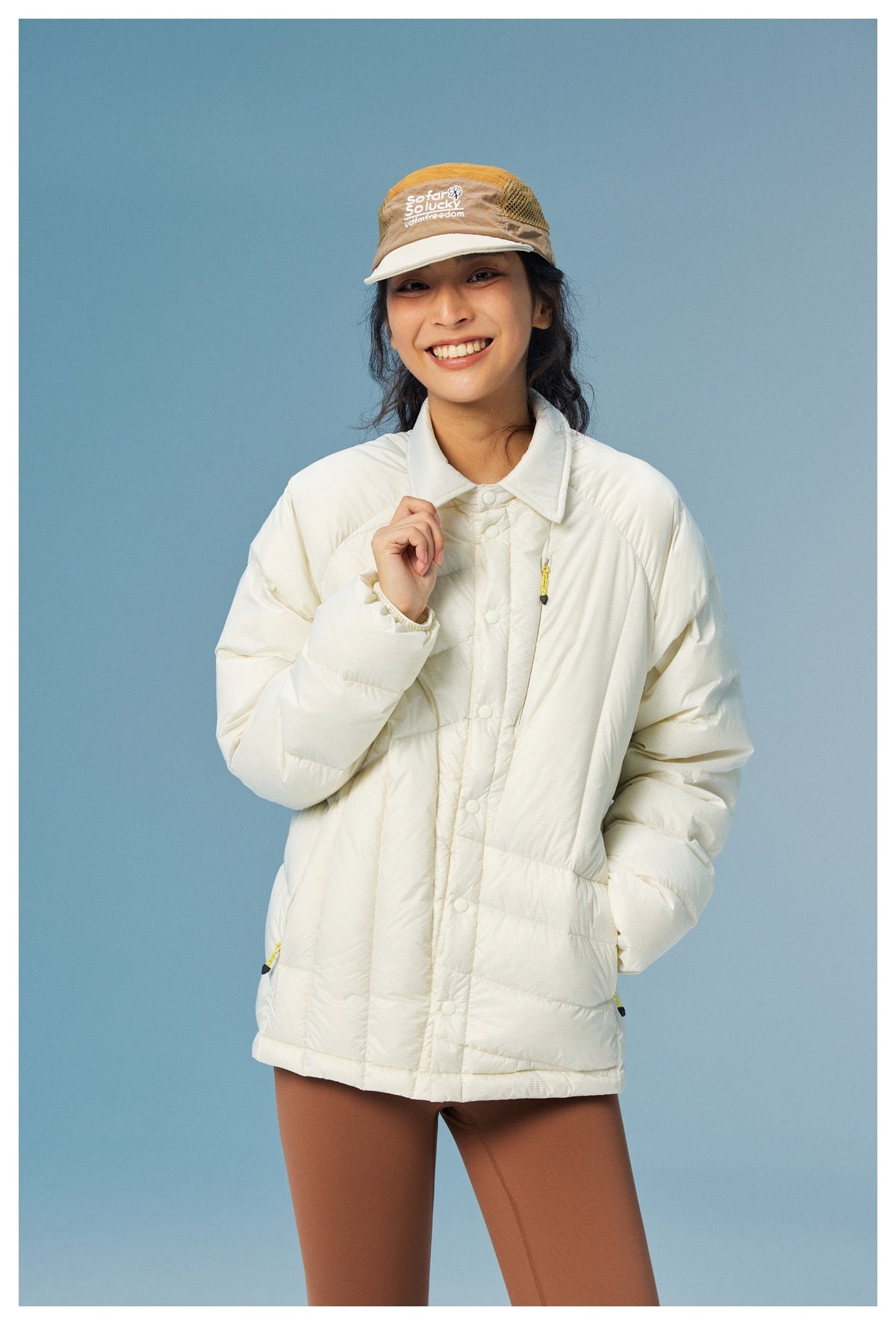 Model wearing a cream collared puffer jacket with snap buttons and yellow zipper accents, paired with brown leggings.