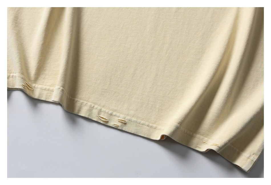 Close-up of the pale cream T-shirt’s bottom hem, showing several small distressed openings and stitching.