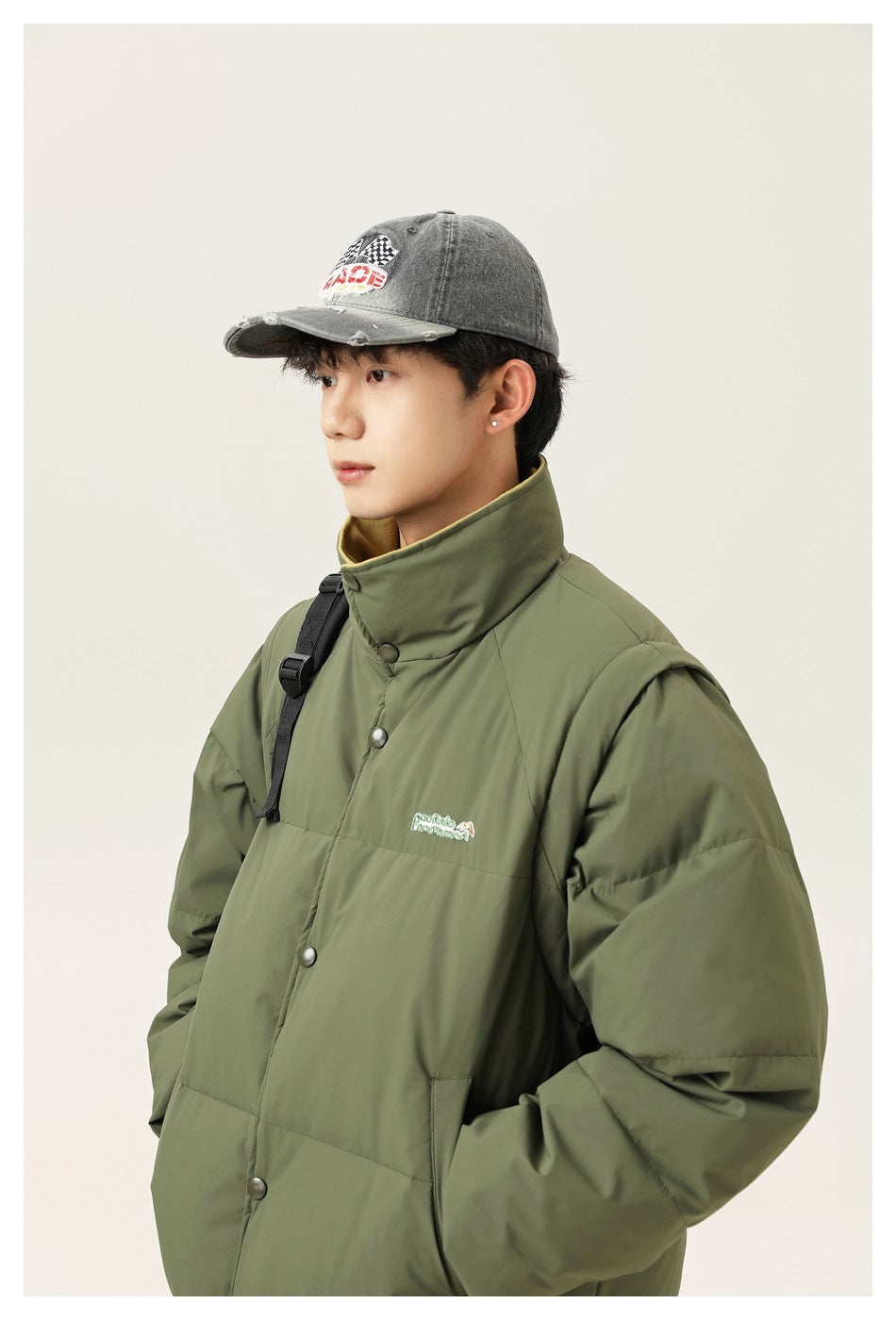A model wears an olive-green padded jacket with a high collar and snap front, paired with a gray graphic cap.