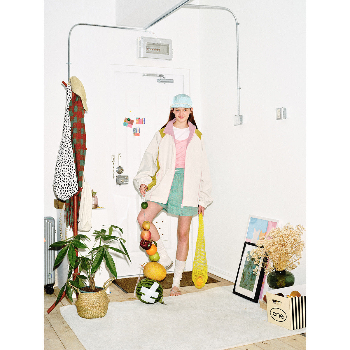 Full-length view of a model in an ivory color-block zip jacket, pink top, turquoise shorts and pale blue cap.