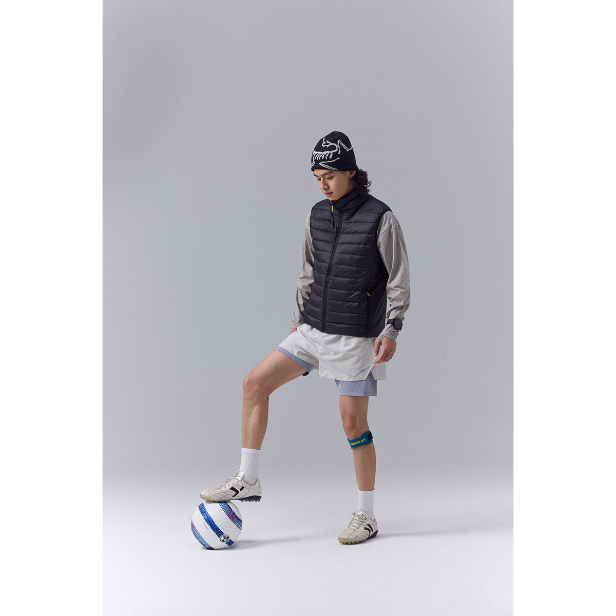 Model wearing a black quilted vest over a gray long-sleeve top, with light shorts and white socks.