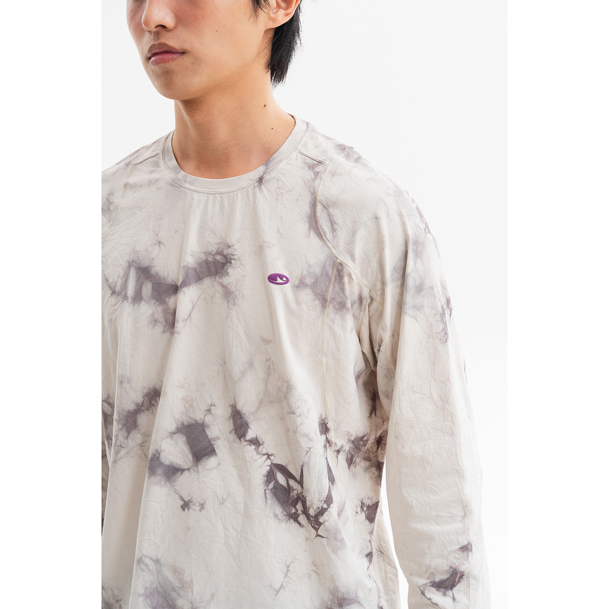Close view of a cream long-sleeve crewneck shirt with gray-purple tie-dye and a small chest emblem, worn by a model.