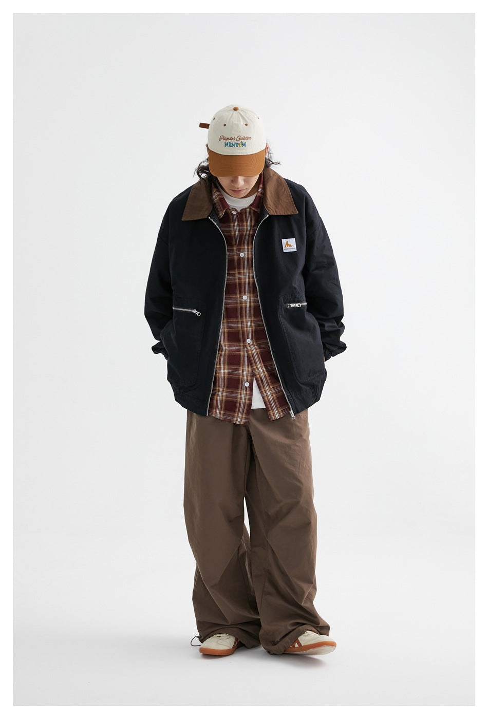 Model with head tilted down wearing the black jacket open over a plaid shirt and brown wide-leg pants