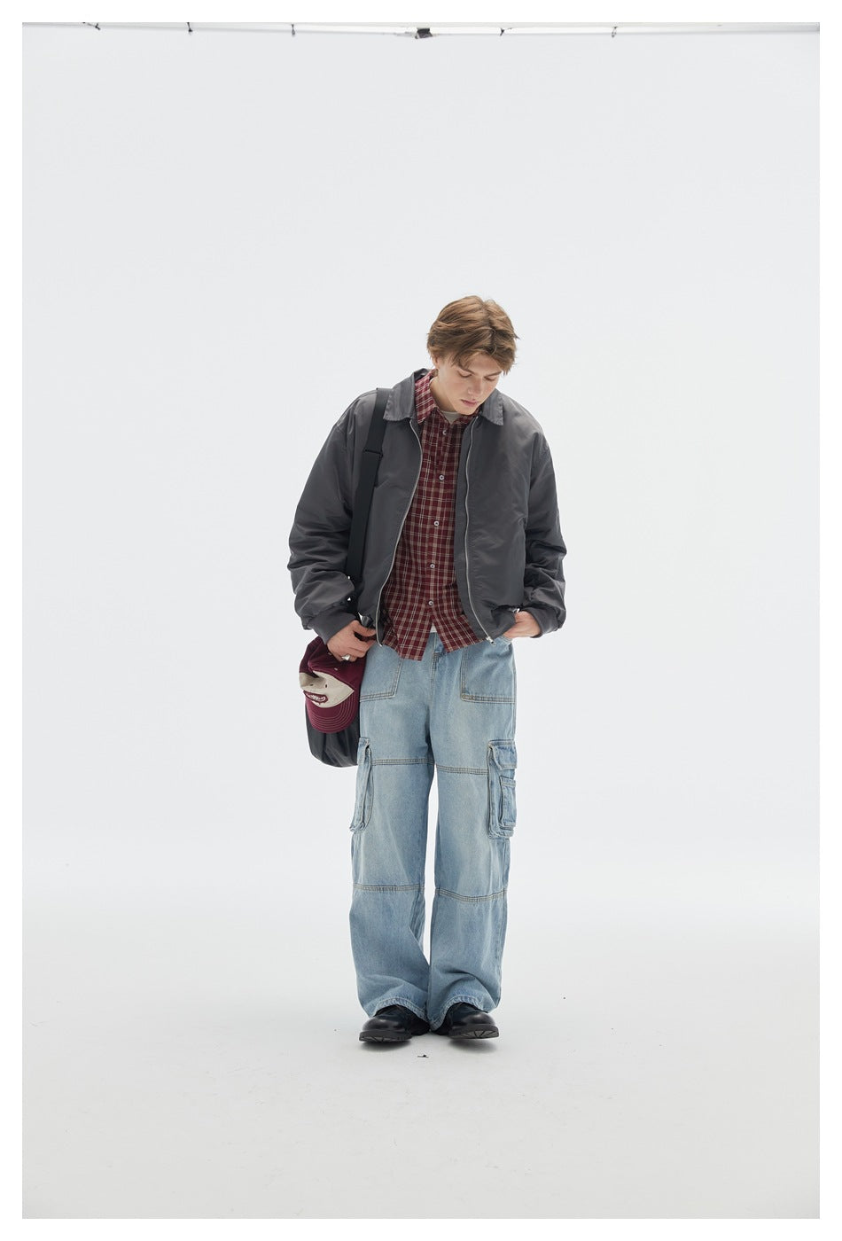 Model wearing a dark gray zip bomber jacket over a plaid shirt with light-wash cargo jeans, looking down with a shoulder bag