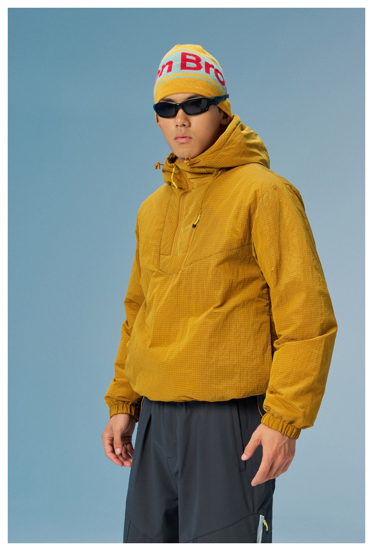 Man wearing a mustard hooded pullover with a subtle grid pattern, dark pants, sunglasses and a yellow knit beanie.