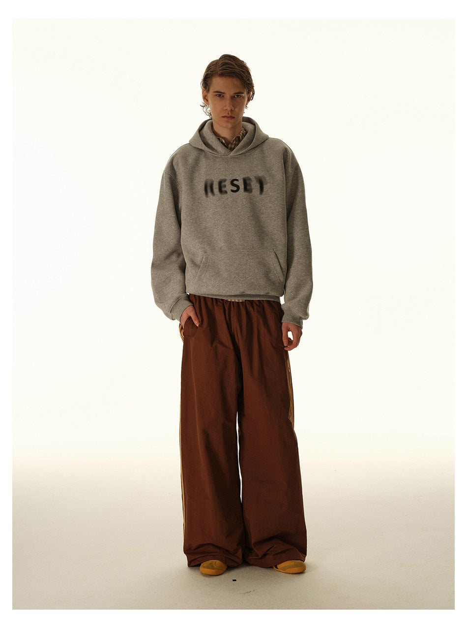 Model wearing a gray hooded sweatshirt with dark chest lettering and loose rust-brown trousers.