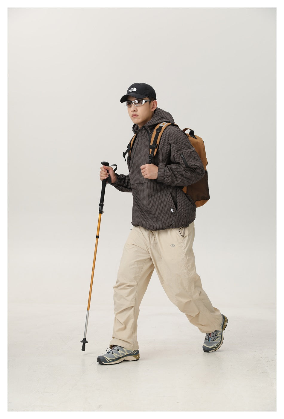 Model walking in a dark brown checked hooded jacket and beige trousers, carrying a backpack and trekking pole.