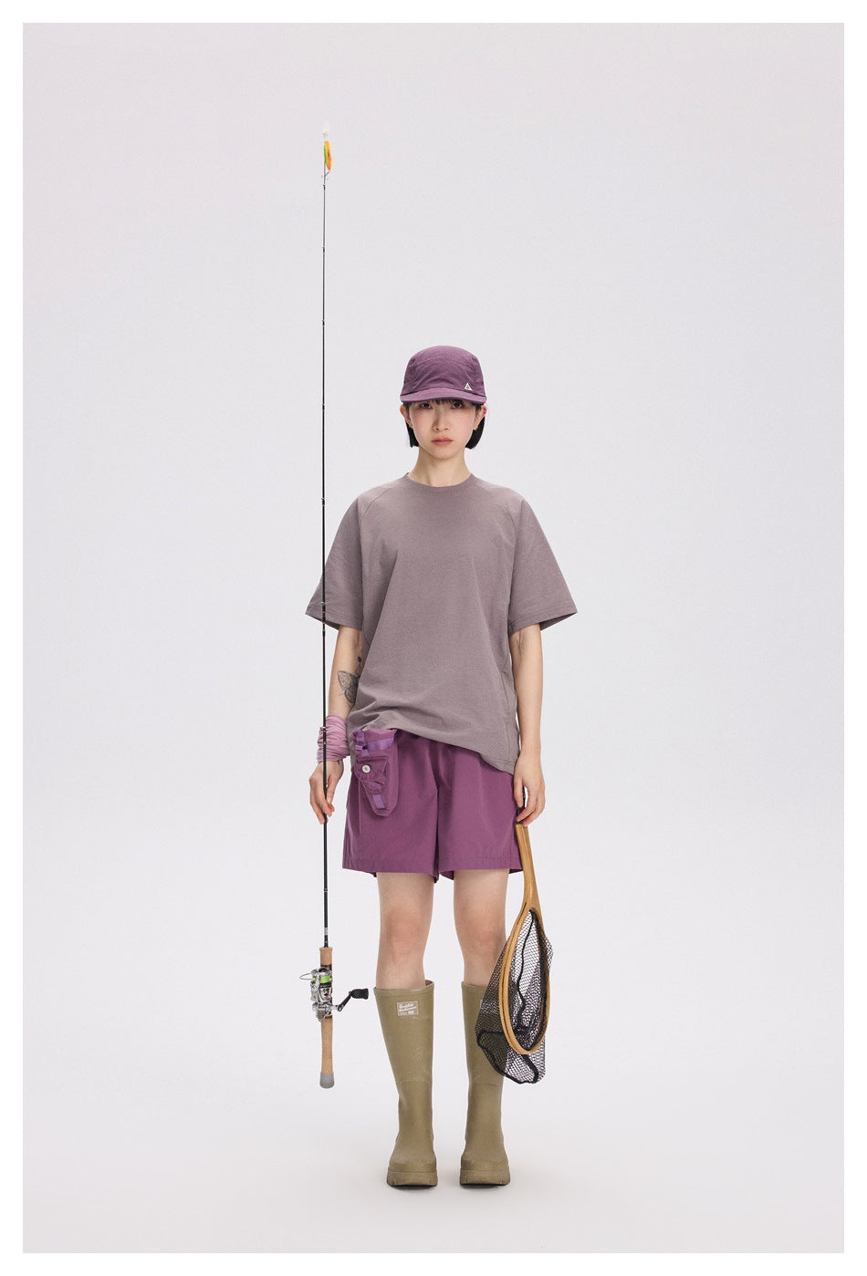 Model wearing a mauve oversized T-shirt, purple shorts, a purple cap, and olive knee-high boots, holding fishing gear.