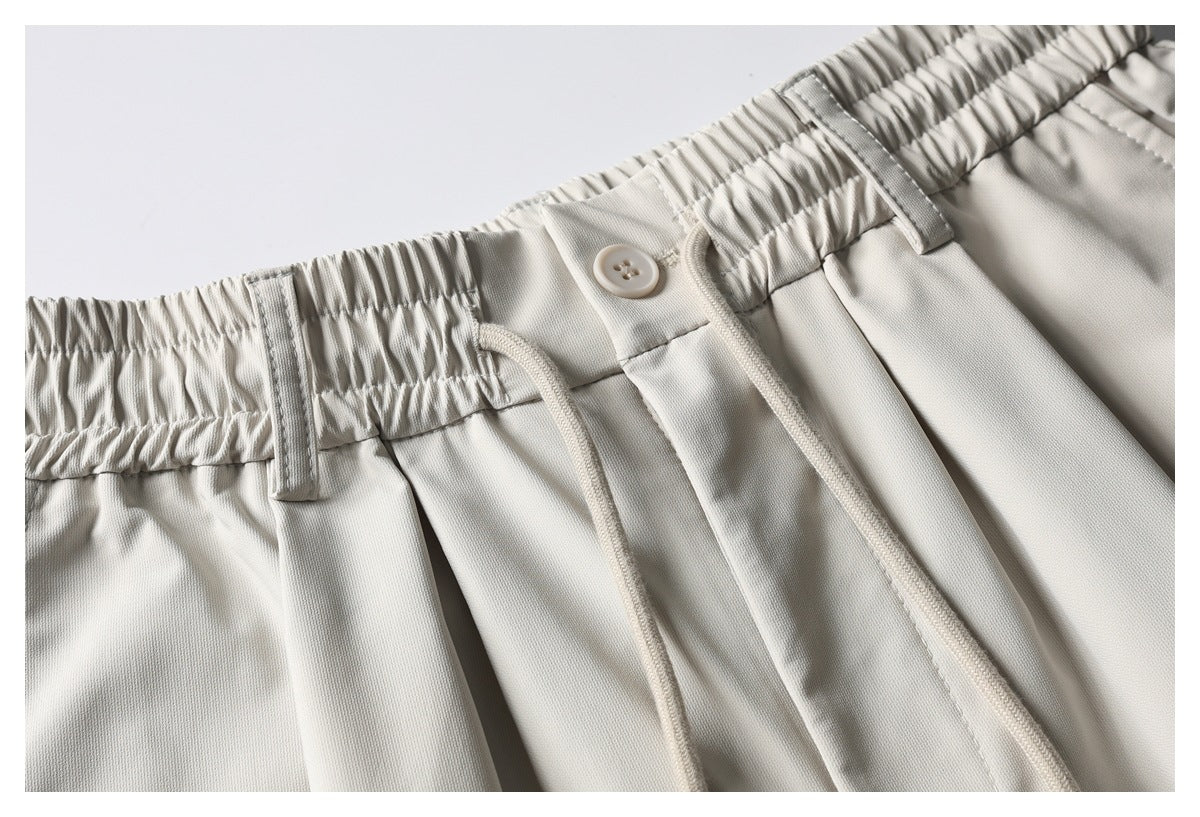 Close-up of light beige shorts showing the elastic waistband, button, belt loops, pleat and drawstrings.