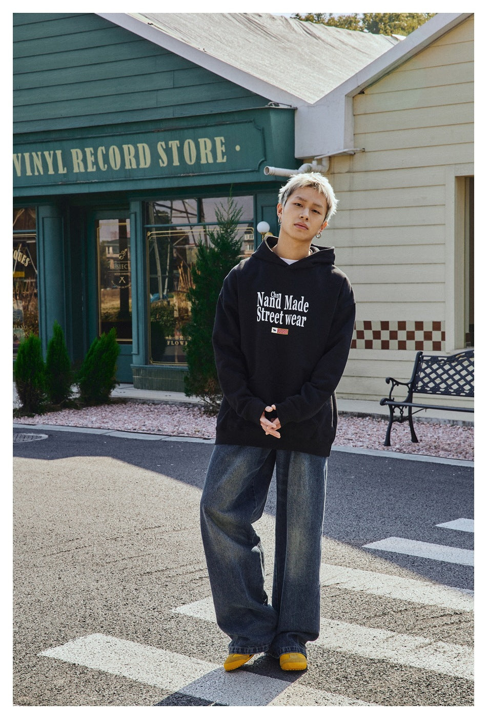 Model wearing a black graphic hoodie with wide-leg faded jeans and yellow shoes, standing in front of a vinyl record store