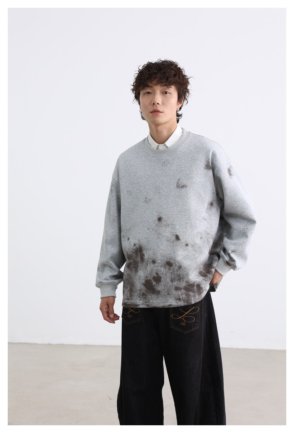 Model wearing a light gray oversized sweatshirt with dark distressed markings concentrated near the hem and black jeans.