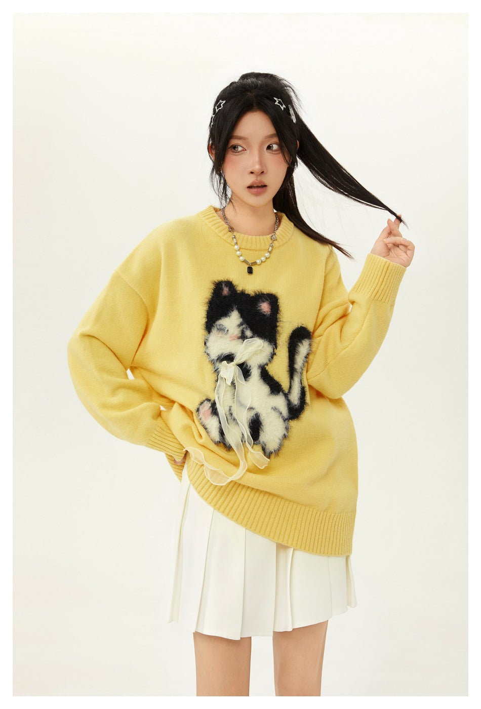 Model wearing a yellow oversized cat-graphic sweater over a white pleated skirt, shown in a playful pose.