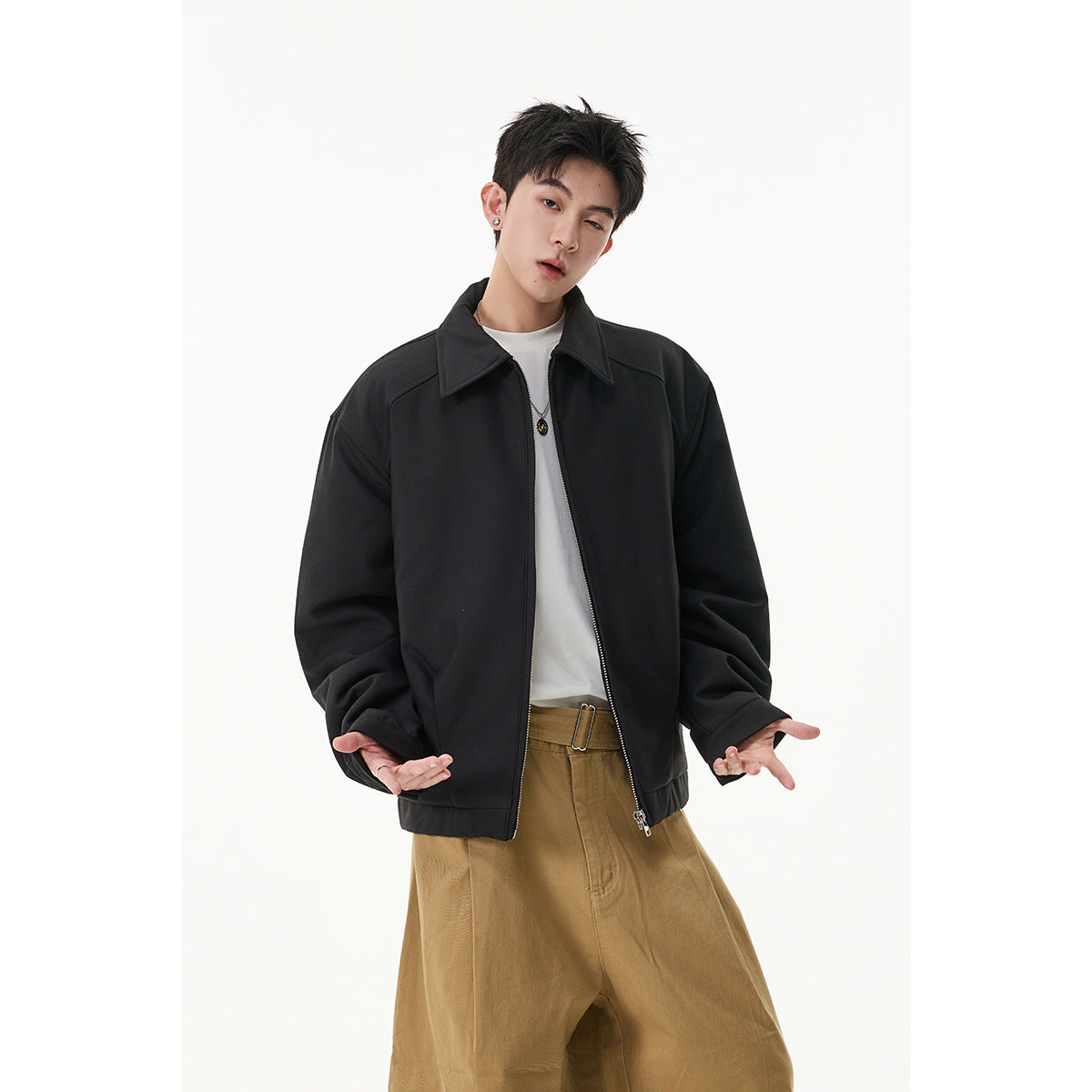 Model wearing a black collared zip jacket open over a white shirt, with loose tan trousers.