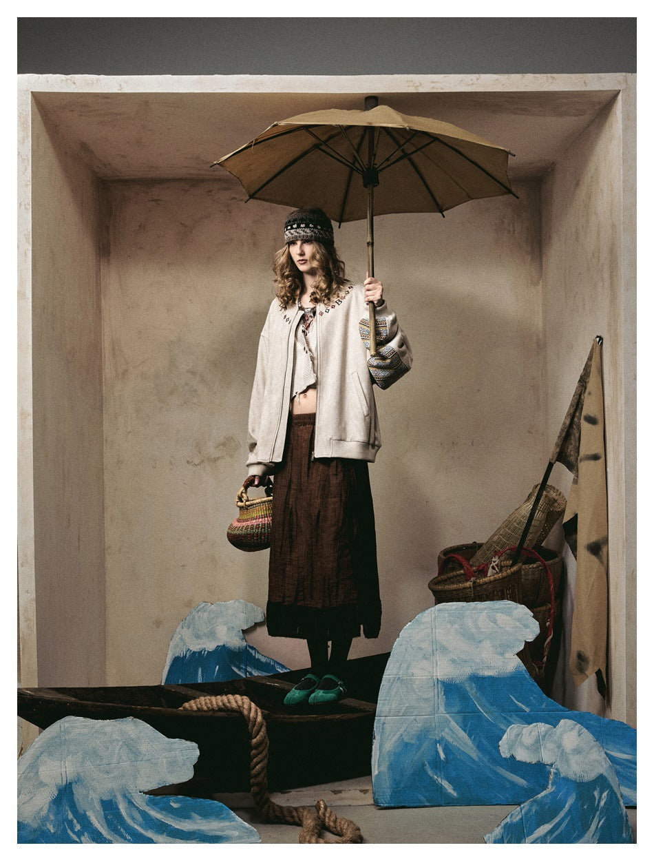 Model in a beige zip bomber jacket turned slightly, showing sleeve trim, holding an umbrella and basket in a styled set