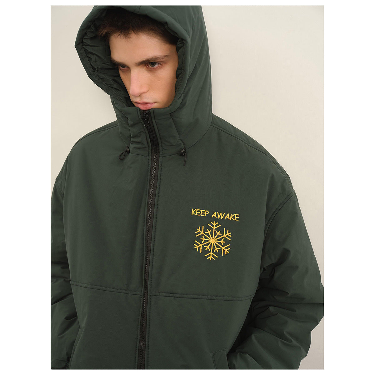 Close view of a model wearing a dark green hooded jacket with a black zipper and yellow snowflake graphic.