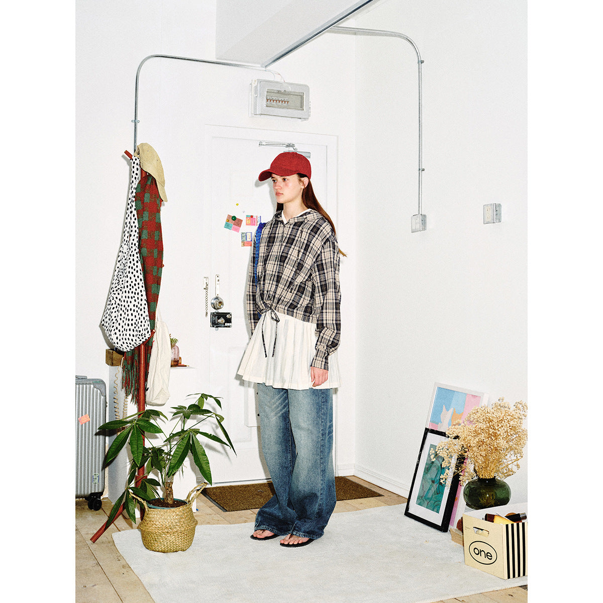 Full-length view of a model wearing a gray plaid hooded button-up with a drawstring hem, white pleated layer, and wide-leg