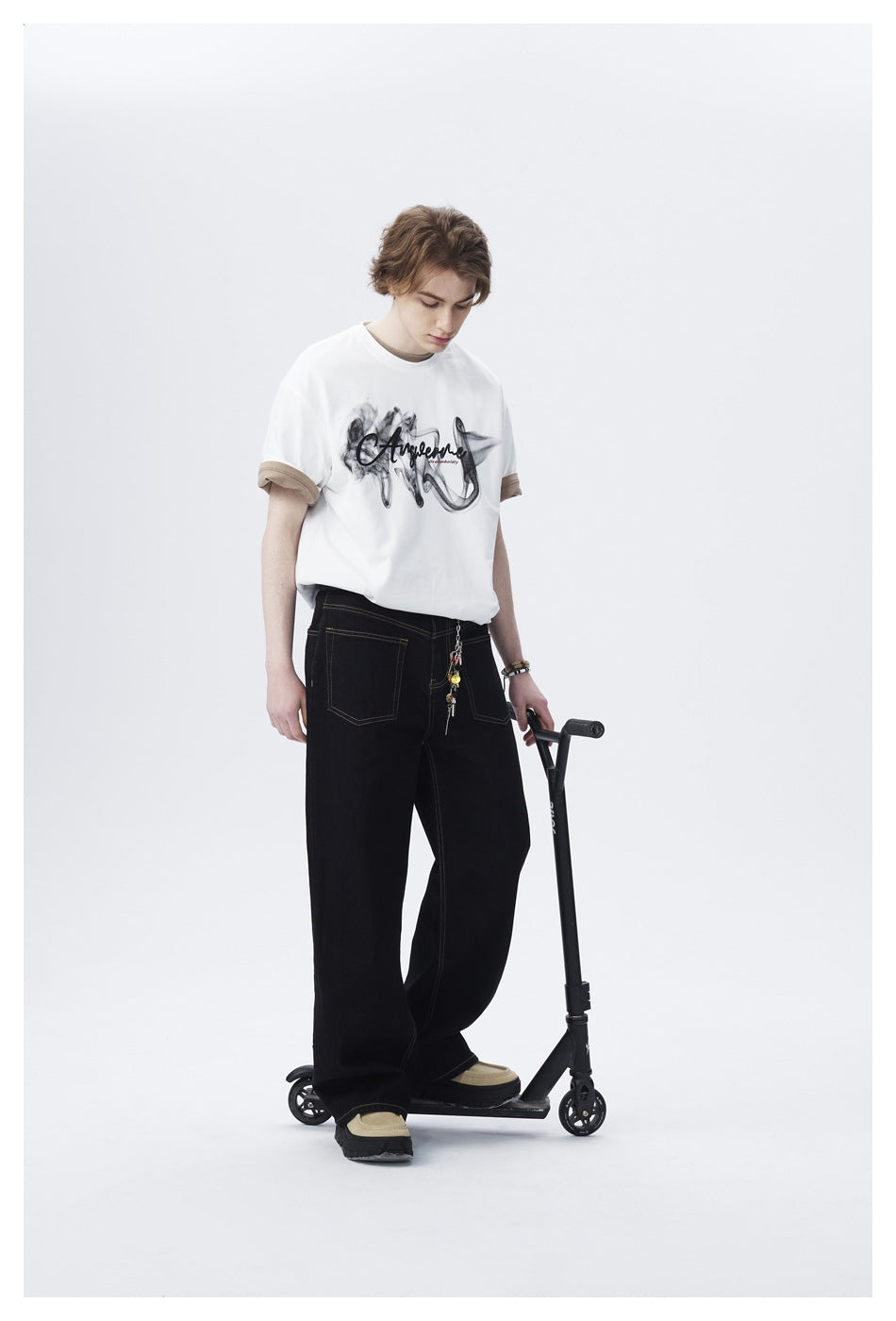 Full-length model wearing a white graphic T-shirt with tan rolled cuffs and loose black trousers beside a scooter.