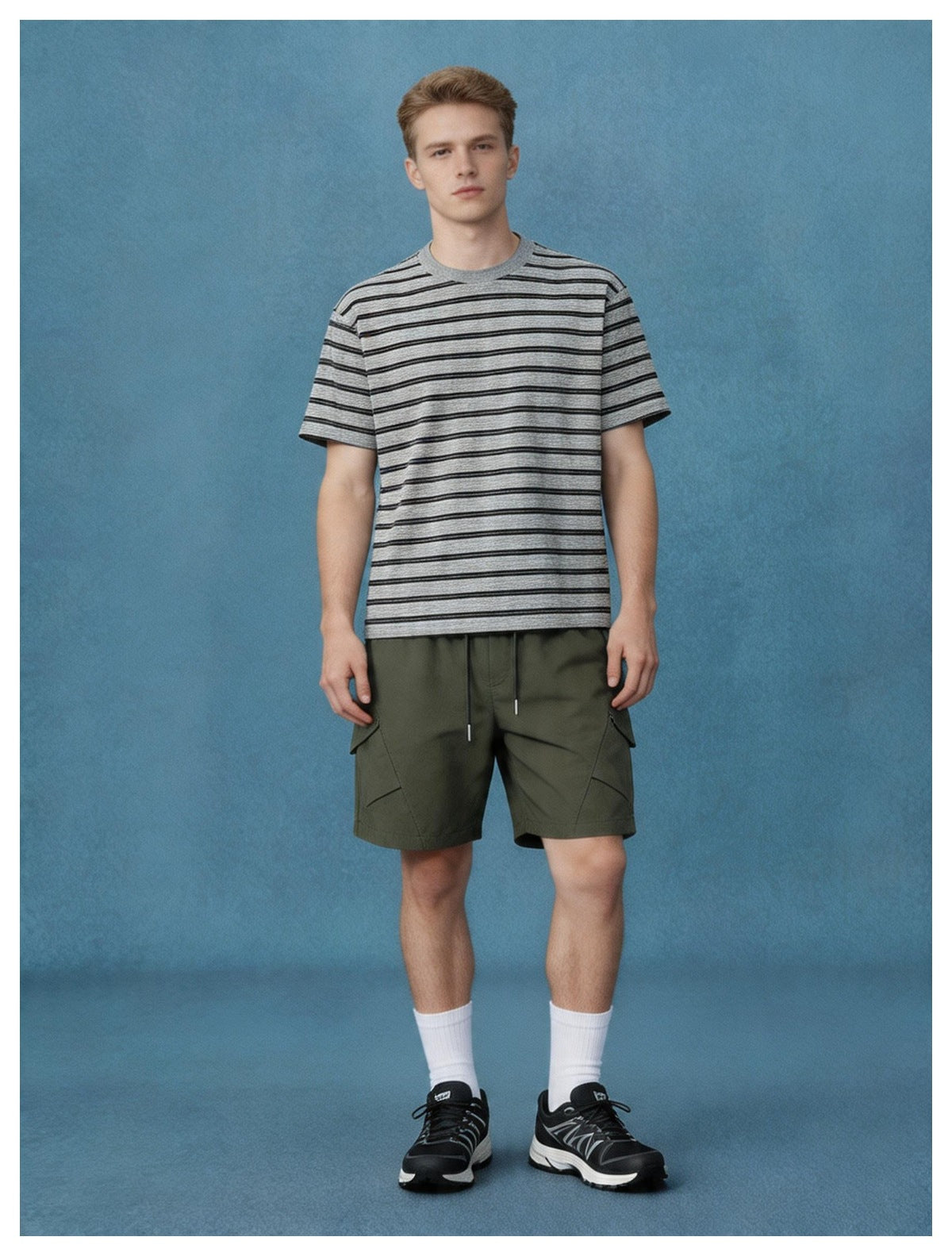 Man wearing army green cargo shorts with a gray striped t-shirt, standing in front of a blue backdrop