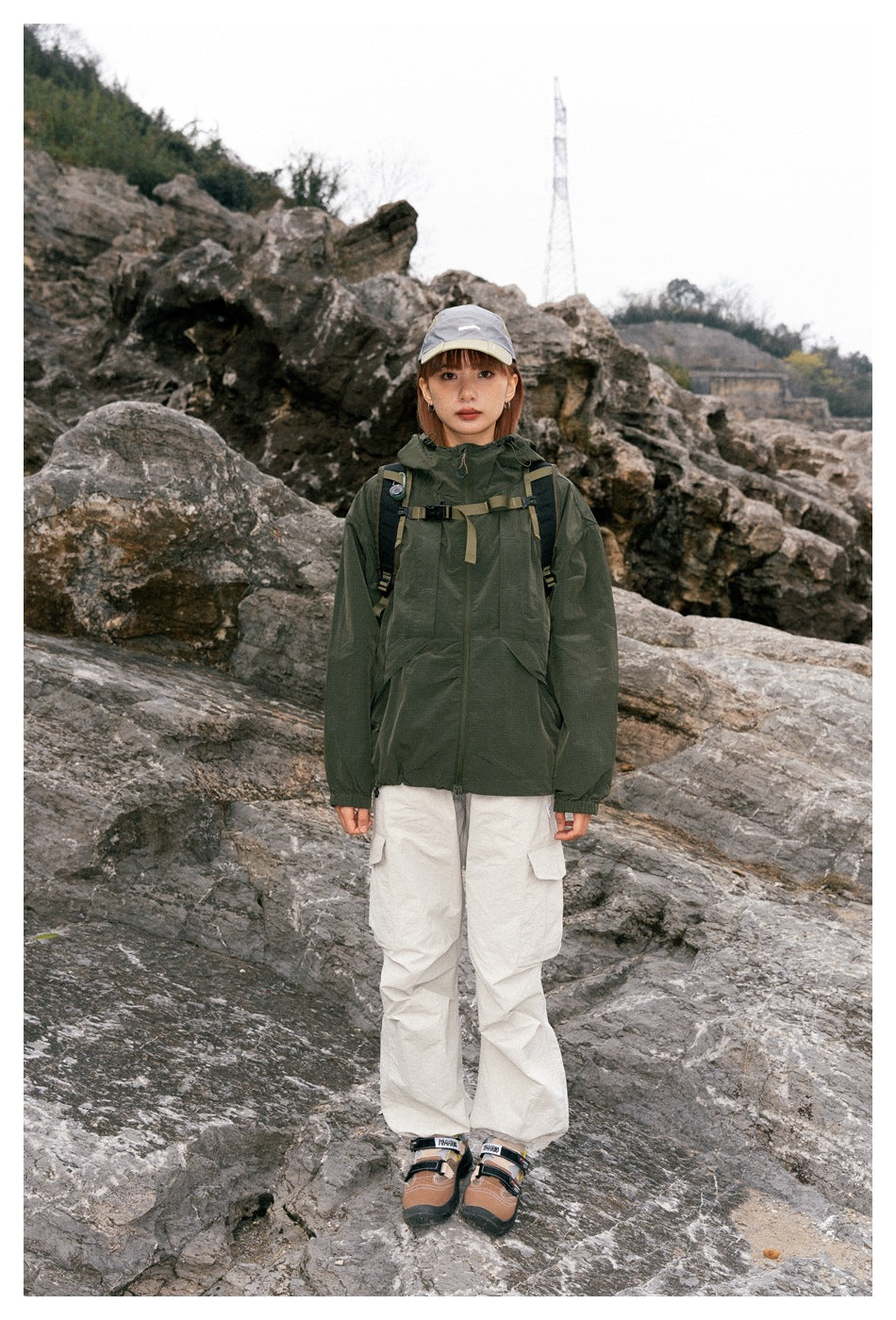 A woman faces the camera in a dark green hooded jacket, off-white cargo pants, and a gray cap.