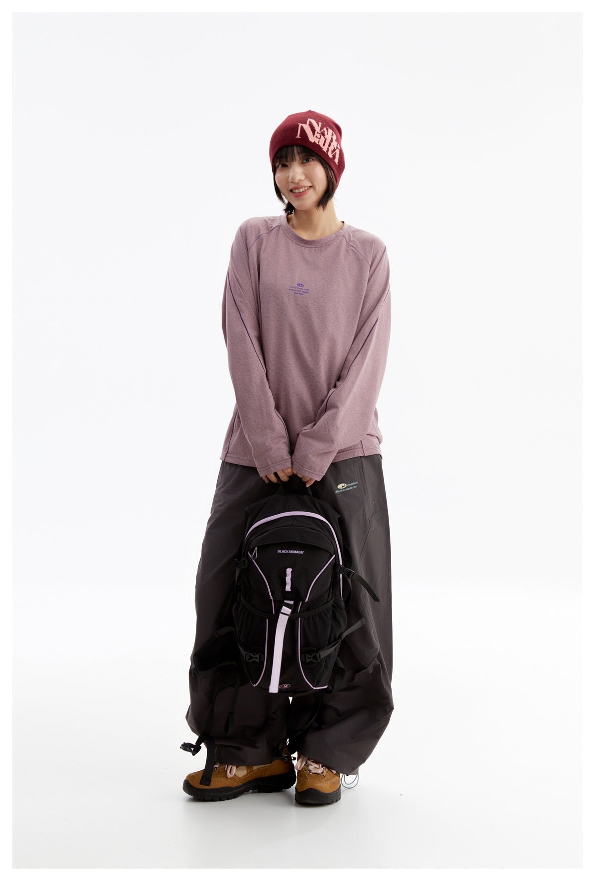Model wearing a mauve long-sleeve top, loose dark pants, and burgundy beanie, holding a black backpack with lilac trim.