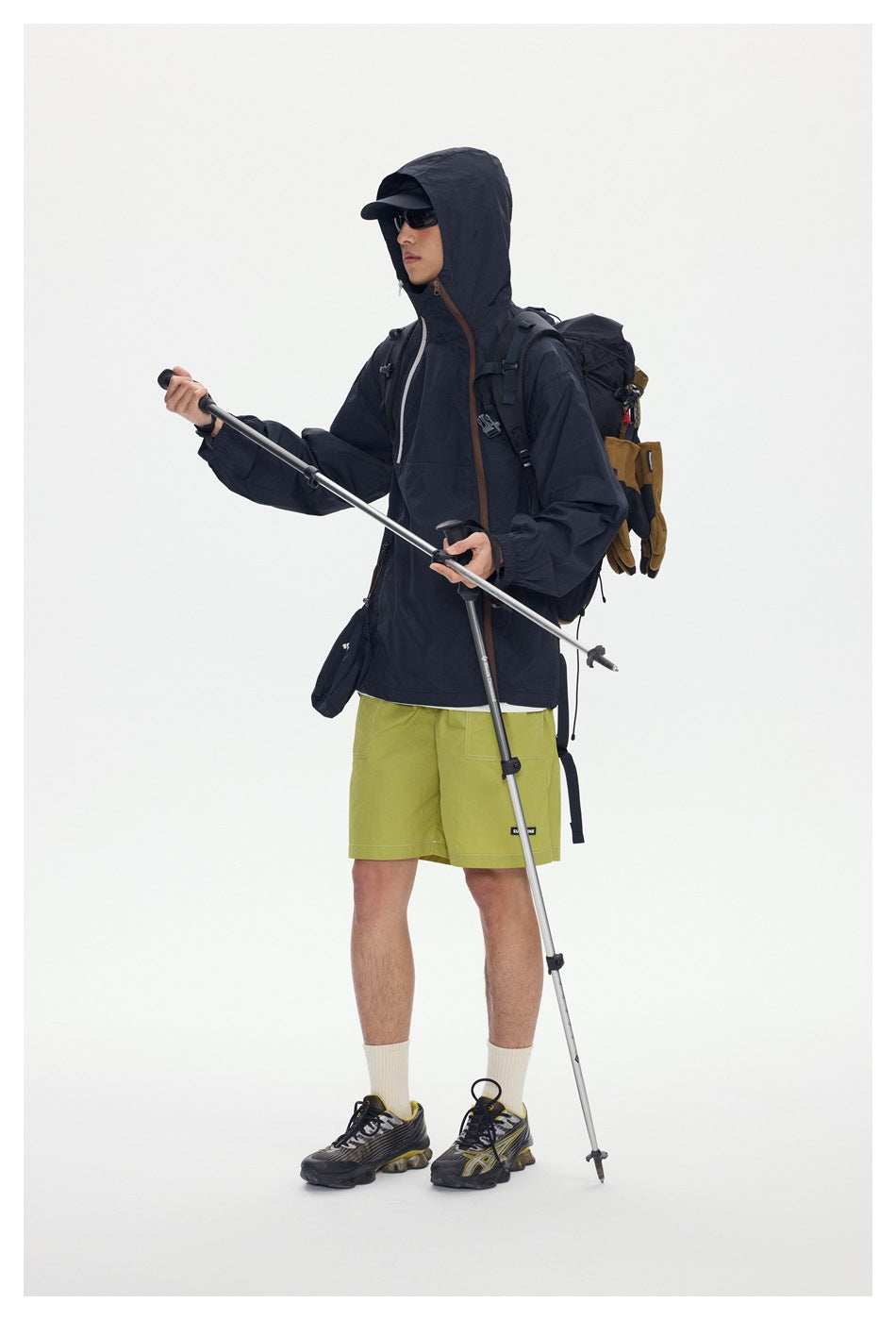 Model in a dark hooded jacket and lime-green shorts, wearing sunglasses and holding trekking poles.