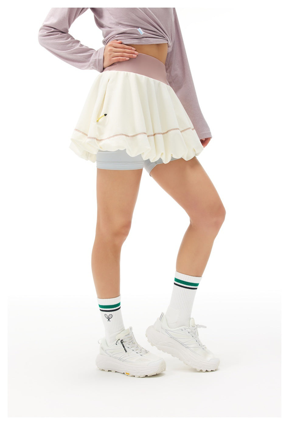 Angled view of an ivory bubble tennis skirt with a mauve waistband over pale gray shorts, worn with a lavender top.