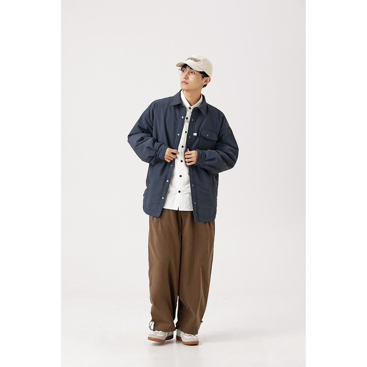 Model wearing a navy snap-front jacket with a chest flap pocket, styled with a white shirt and loose brown trousers.