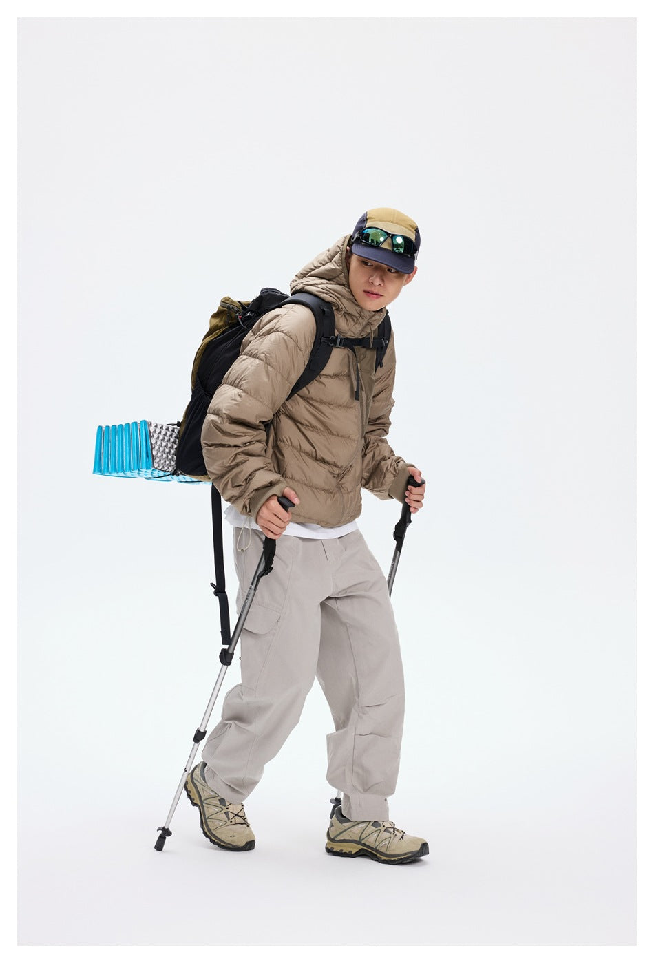 Model wearing a taupe hooded puffer jacket and light gray trousers with a hiking backpack and trekking poles.
