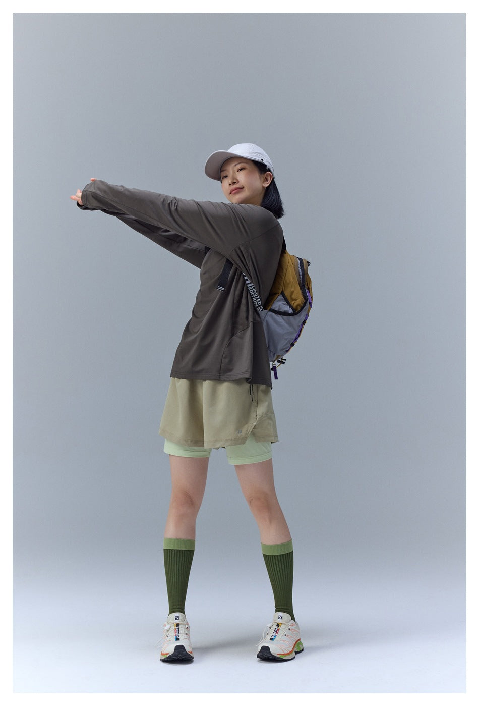 Model in a dark taupe long-sleeve quarter-zip top and layered khaki and pale green shorts, stretching outdoors.