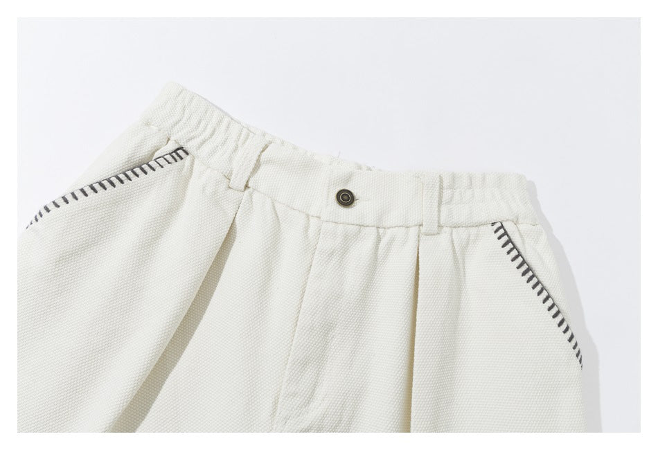 Cropped front view of ivory shorts showing the buttoned waistband, pleats and dark contrast stitching around the pockets.