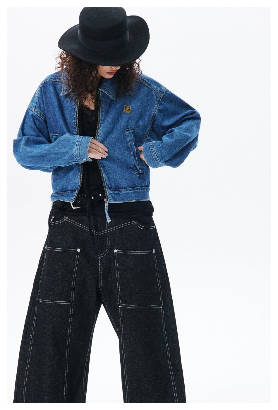 Close crop of model wearing blue denim zip jacket with star patch, hat covering the face, over black wide-leg jeans