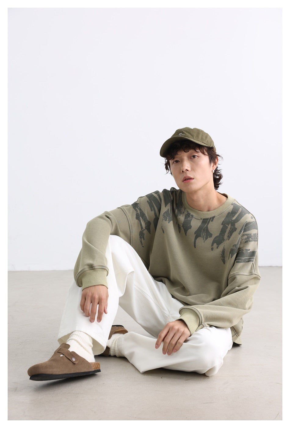 Person seated on the floor wearing the green printed sweatshirt with a cap, white pants and brown clogs, facing front