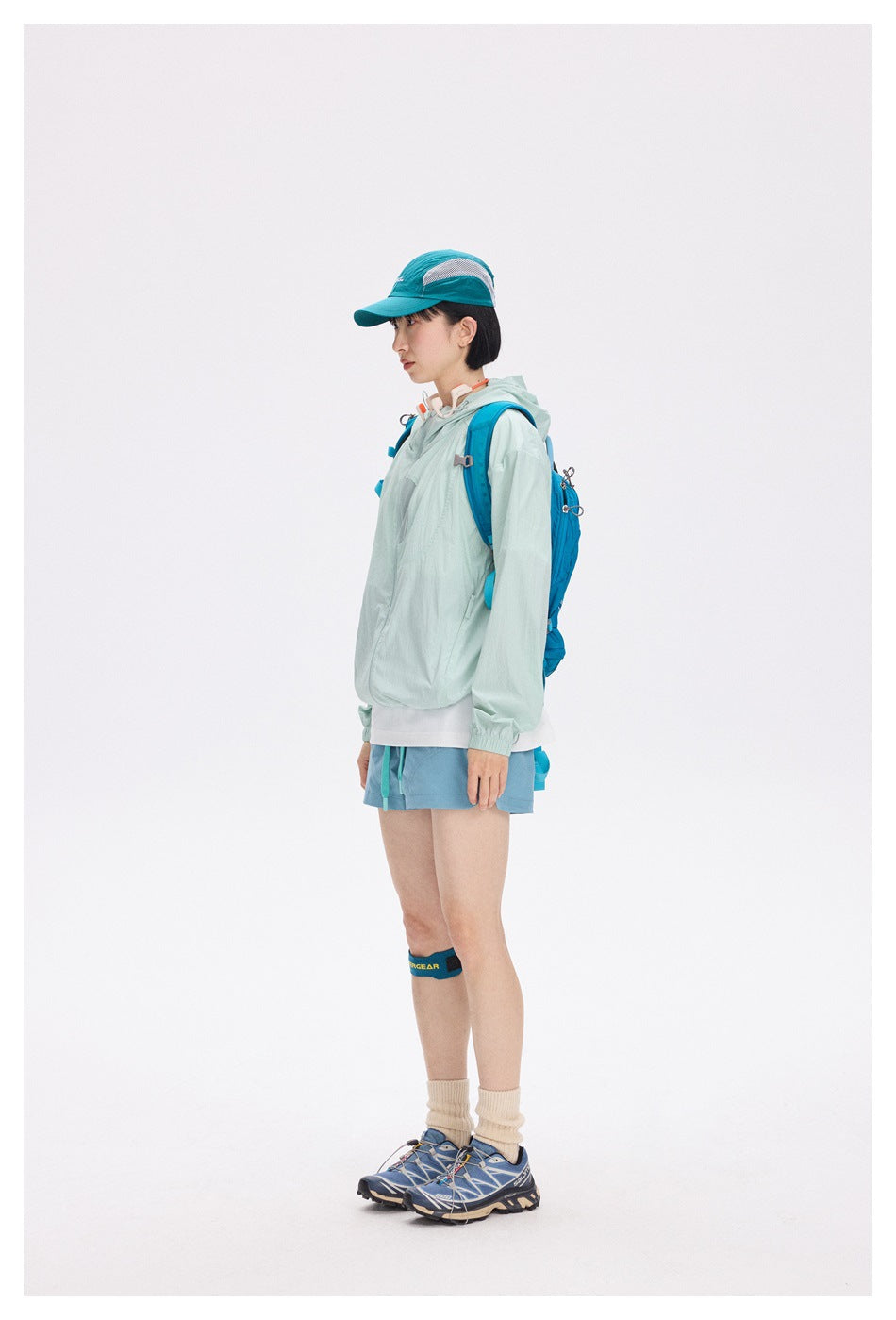 Model in profile wearing a pale mint hooded jacket, blue shorts, and a blue hiking backpack.