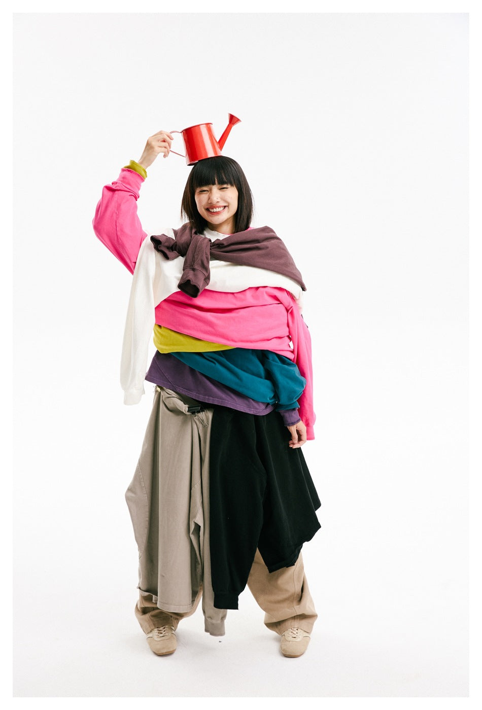 Smiling woman poses in layered colorful tops with a red watering can balanced on her head.