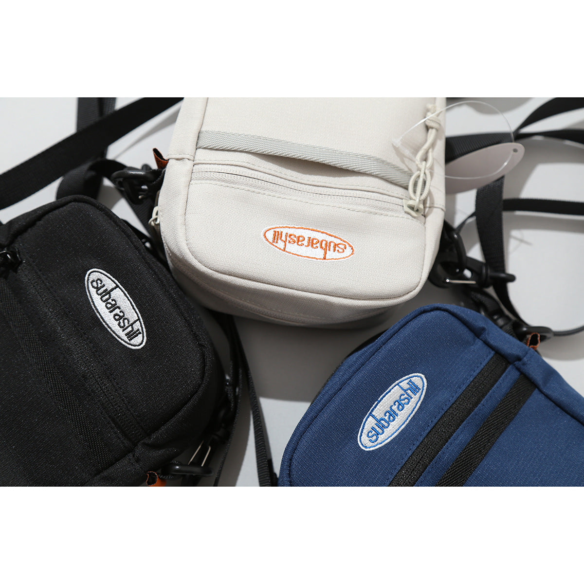 Three small crossbody bags in black, off-white, and navy, with adjustable black straps and front zippers.