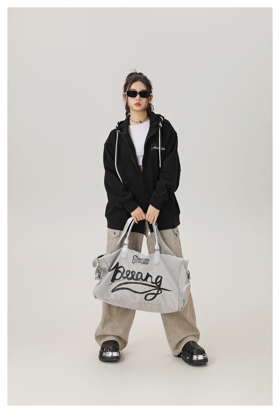 Model wearing an oversized black zip-up hoodie with white drawstrings, holding a large gray printed tote bag.