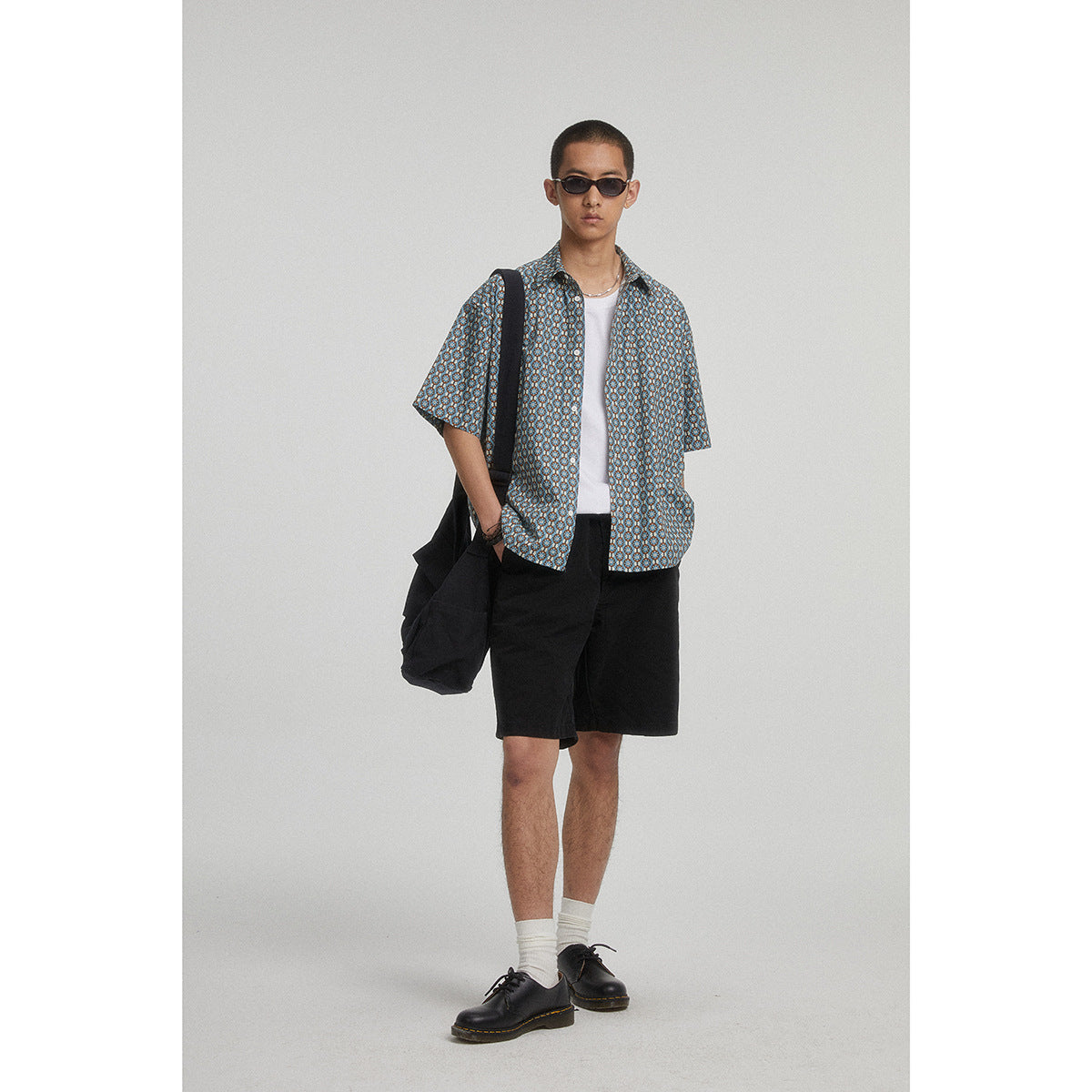 Model wearing a blue geometric-pattern short-sleeve button-up over a white tee with black shorts.