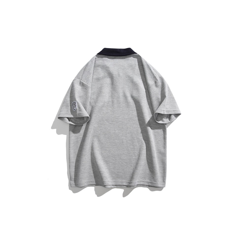 Back view of a light gray short-sleeve polo with a dark collar and a small shield patch on the sleeve.