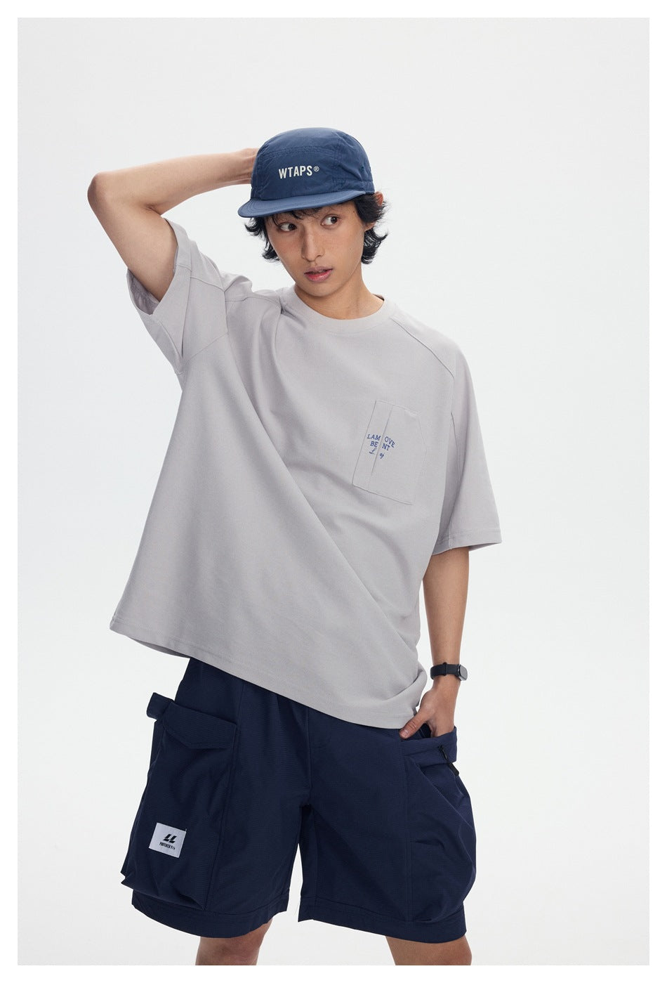 Model wearing an oversized light gray pocket T-shirt, navy cargo shorts, and a navy cap.