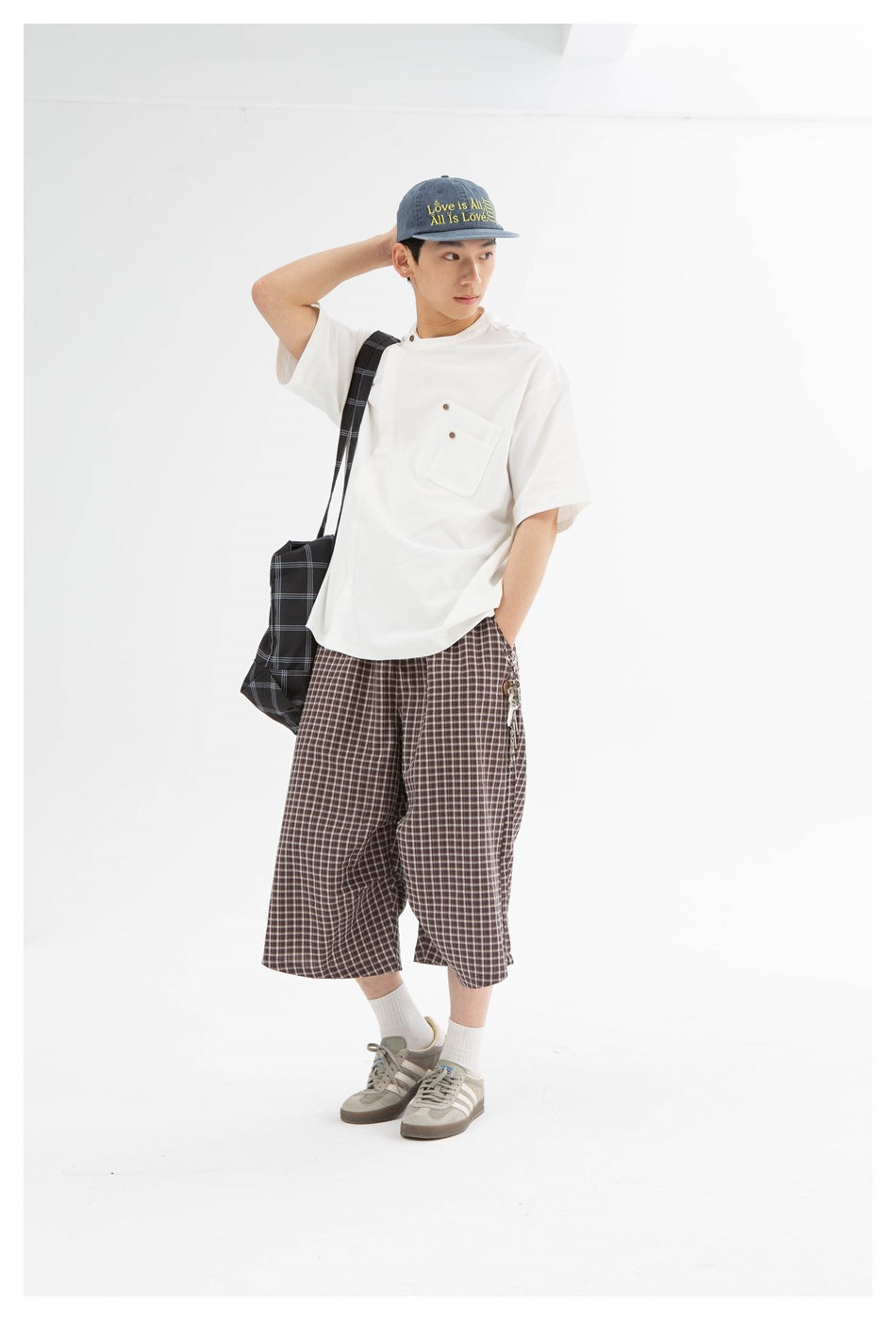 Man in a white relaxed short-sleeve tee with a buttoned chest pocket, checked pants, cap and sneakers