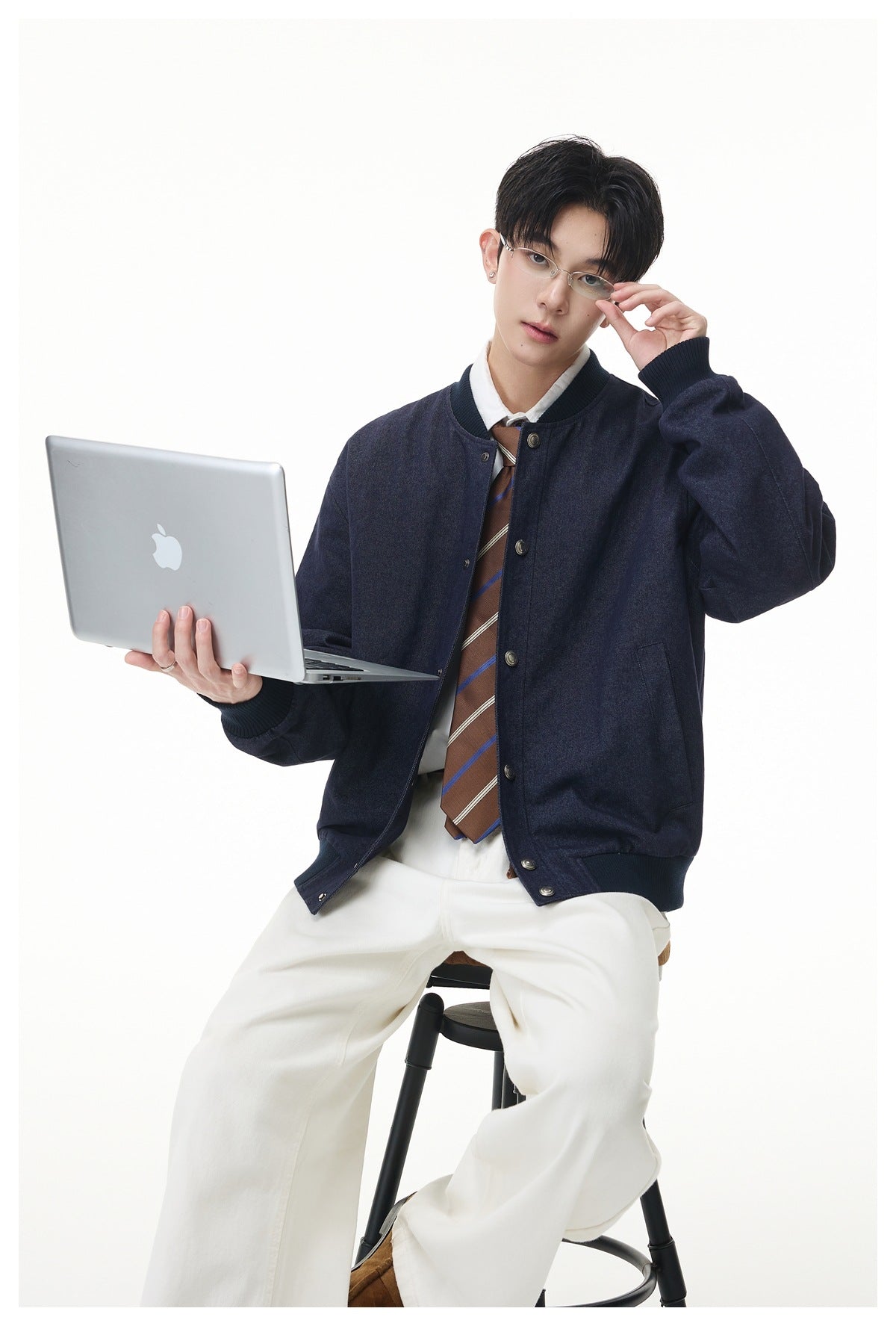 Model seated on a stool wearing a navy snap-front jacket with a striped tie and white pants, holding a laptop