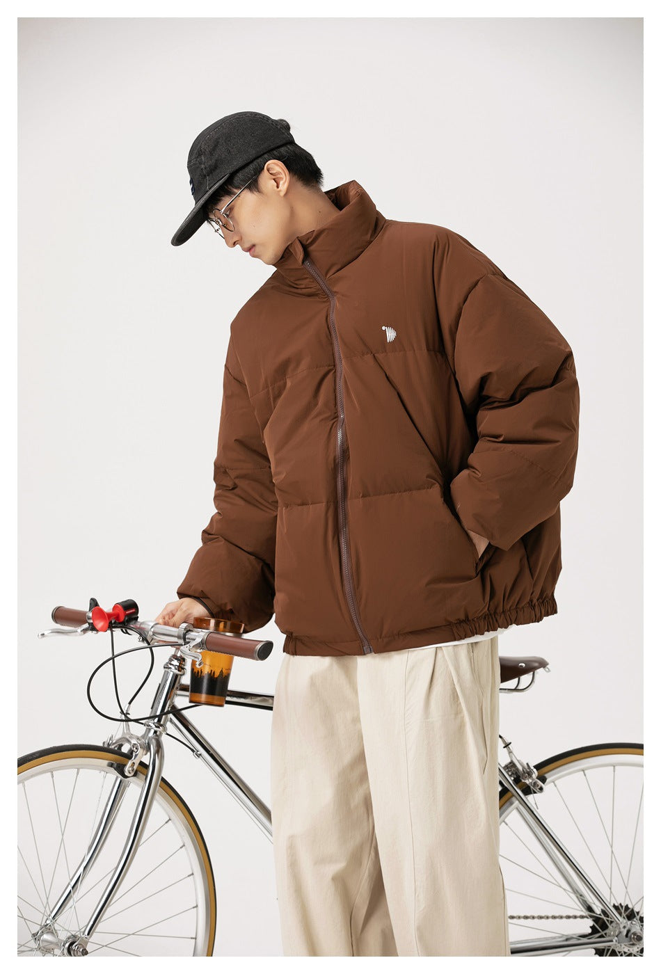 Model wearing a brown quilted zip-up jacket with a high collar and small chest emblem beside a bicycle.