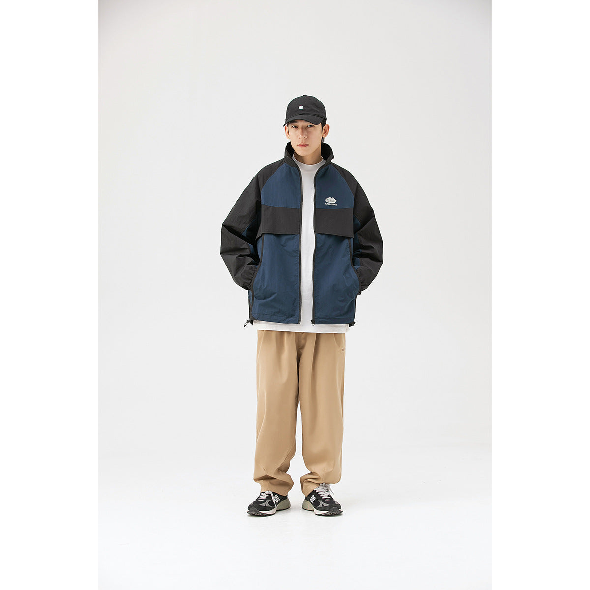 Full-length front view of a model wearing a navy and black color-block zip jacket with beige trousers.