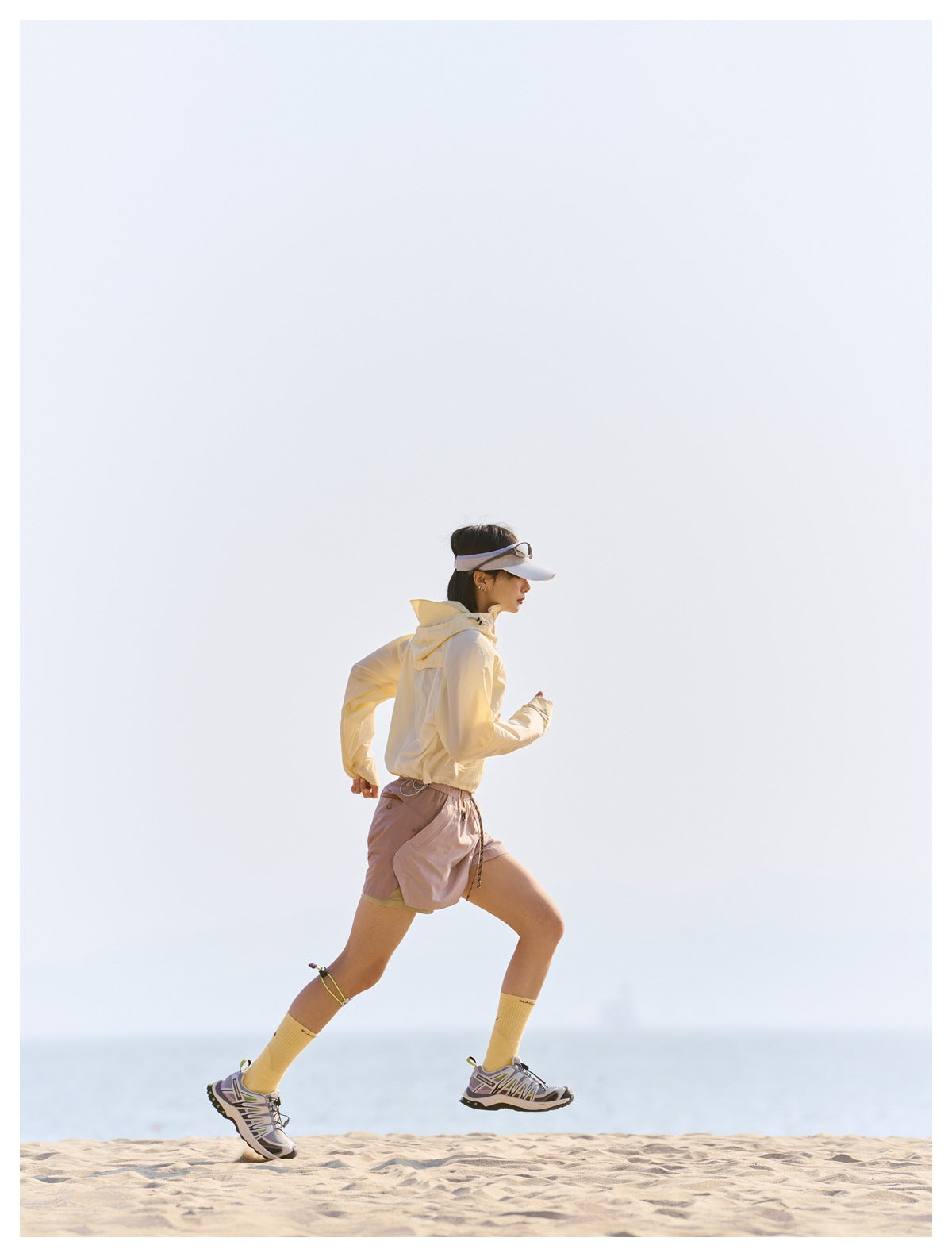 A woman runs along a beach wearing a pale yellow hooded jacket and dusty pink athletic shorts.