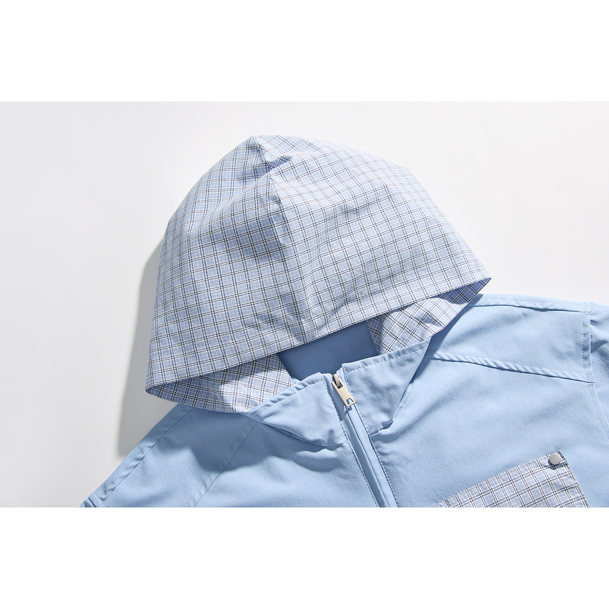Close-up of a light blue jacket with a blue plaid hood, front zipper, and matching plaid pocket.