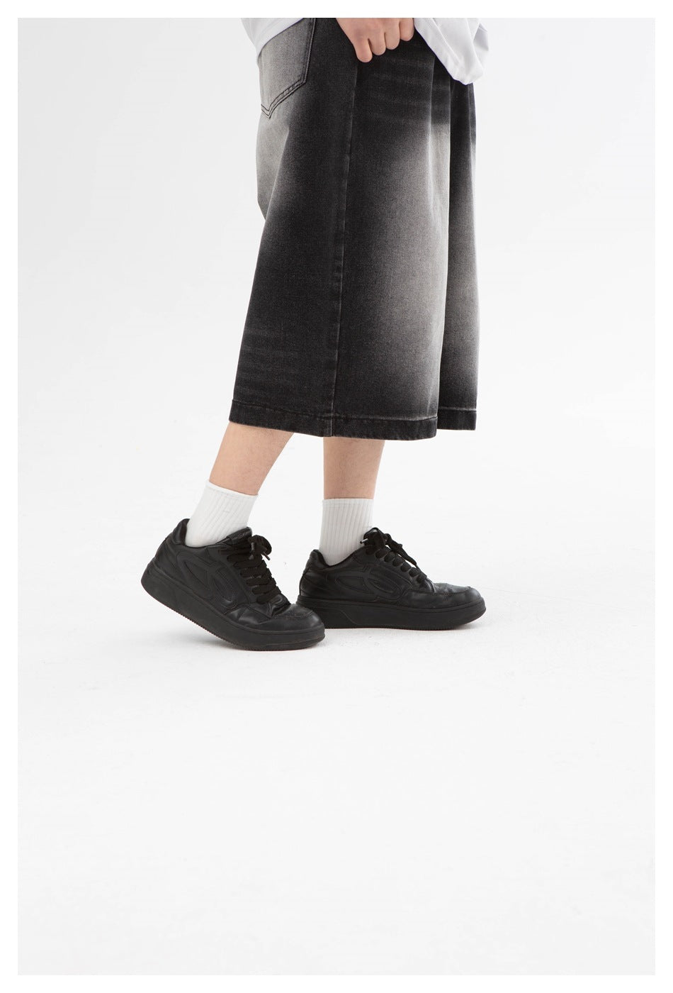 Model wearing wide-leg, below-knee charcoal-black denim shorts with faded panels and black sneakers.