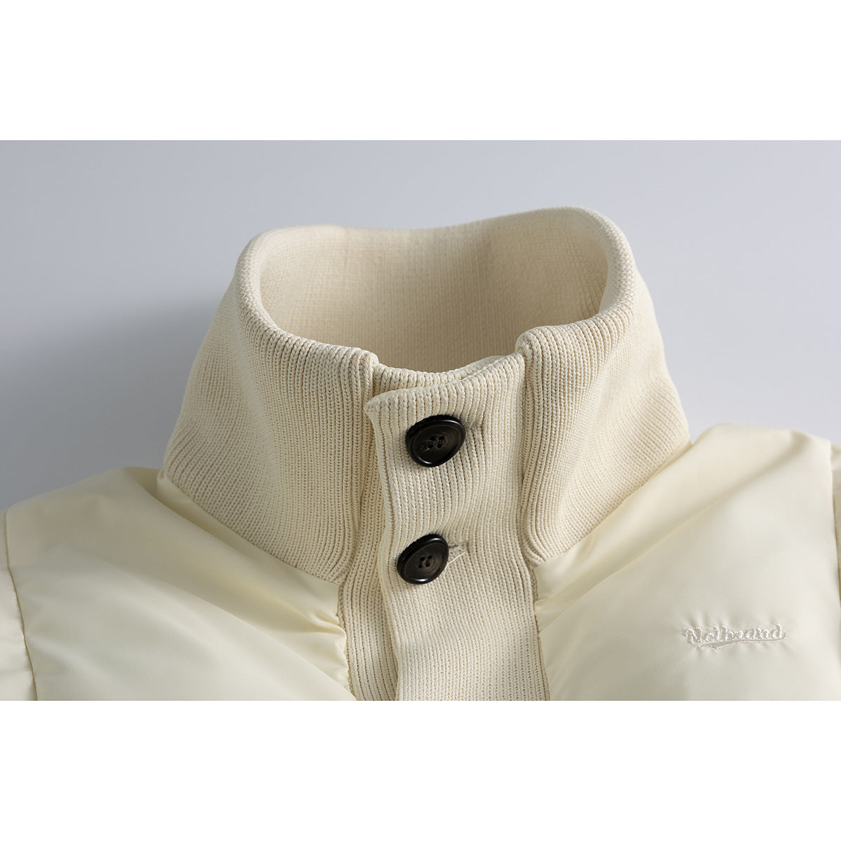 Close-up of an ivory padded jacket's tall ribbed knit collar and placket with two dark buttons.