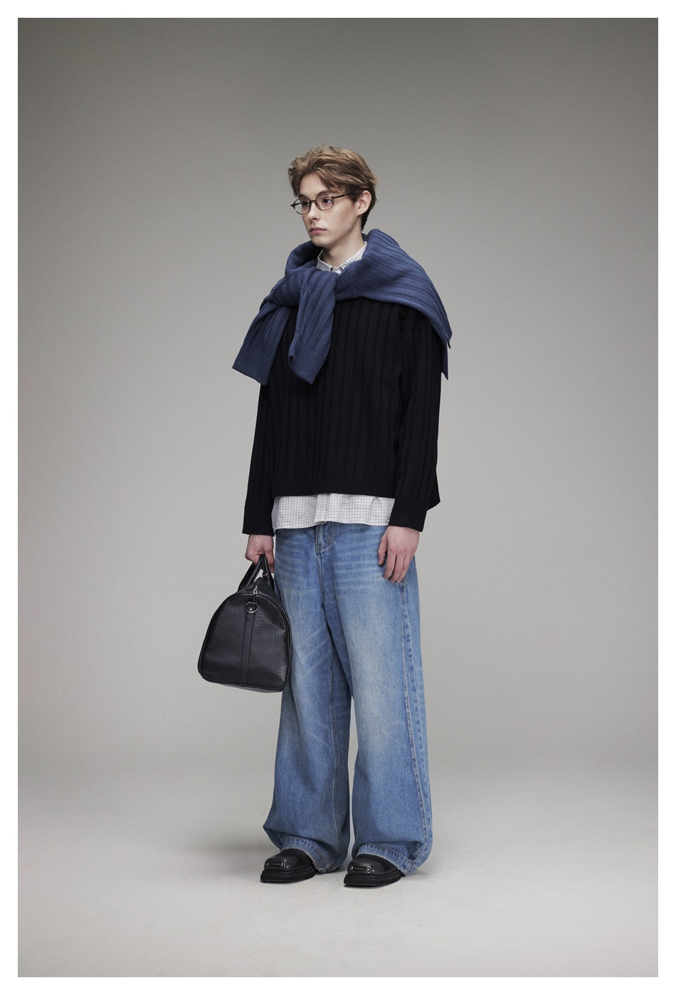 Model wearing a black ribbed sweater over a checked shirt, with a blue sweater draped over his shoulders and wide-leg jeans.